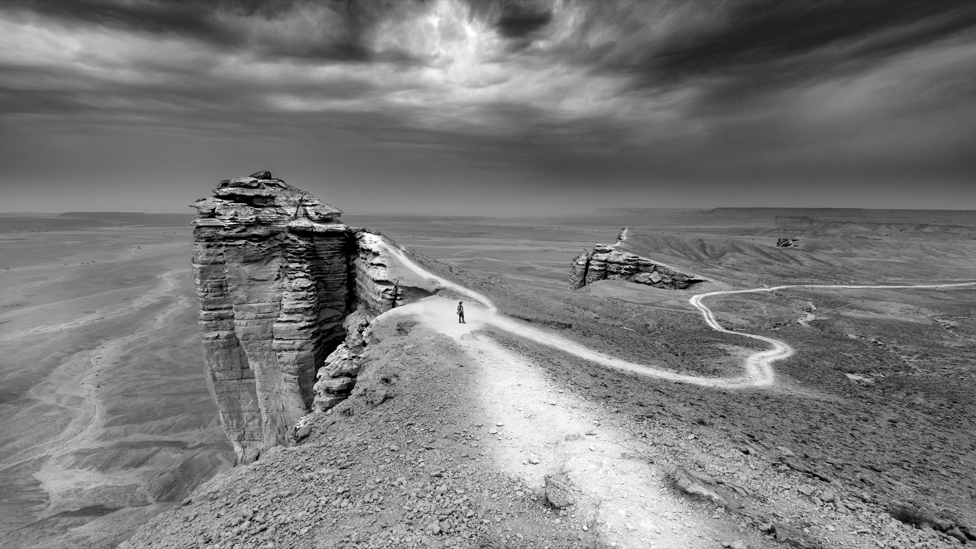 Black and white photograph of a lone figure on a desert ridge beneath a dramatic sky